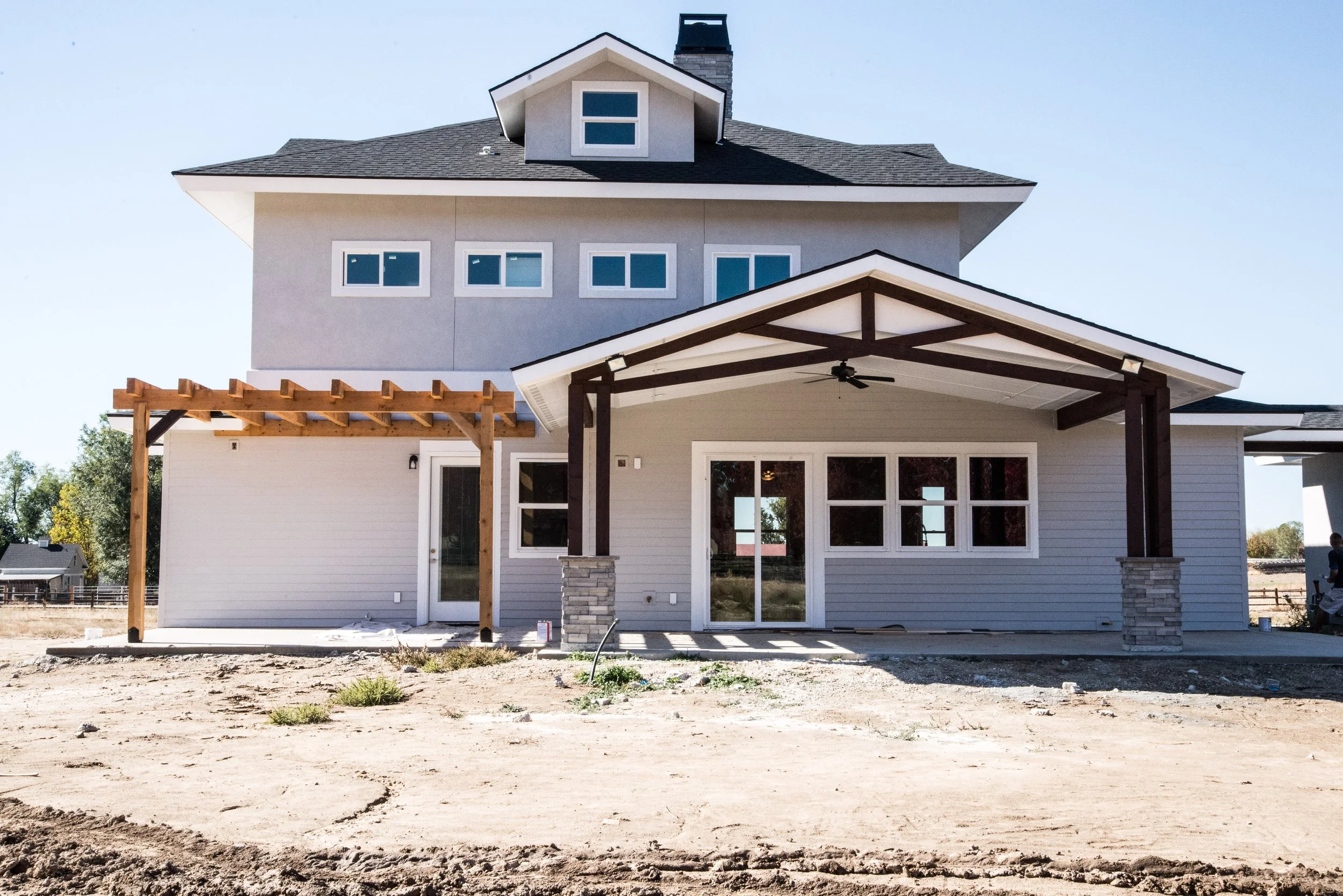 Front view of a two-story house under construction with unfinished yard, featuring a covered porch, multiple windows, and a small dormer on the roof.