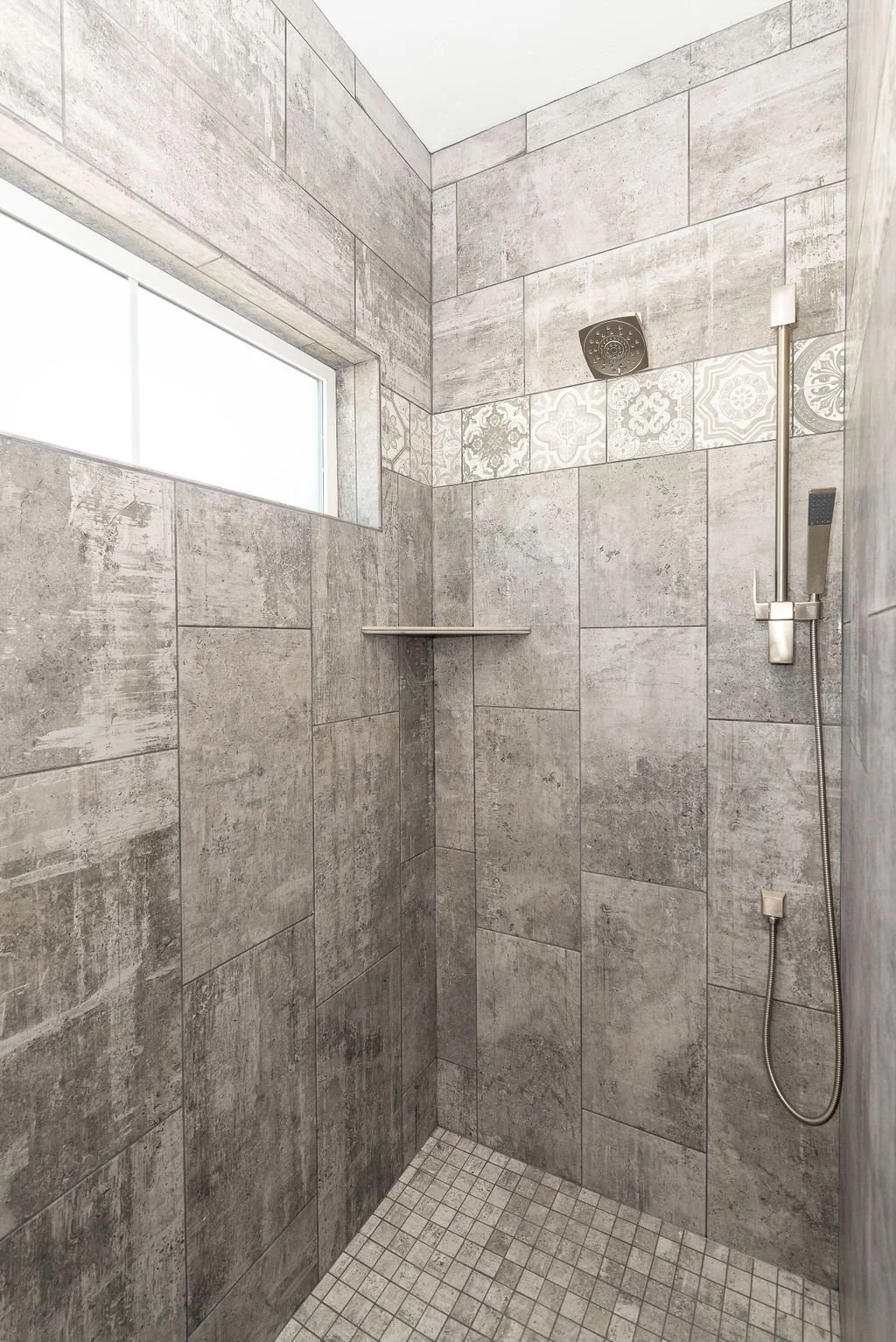 Inside a modern shower with grey textured tile walls and small square tiled floor. There is a window near the ceiling, a black showerhead on the upper wall, a handheld shower wand with a hose on the right wall, and a small corner shelf.