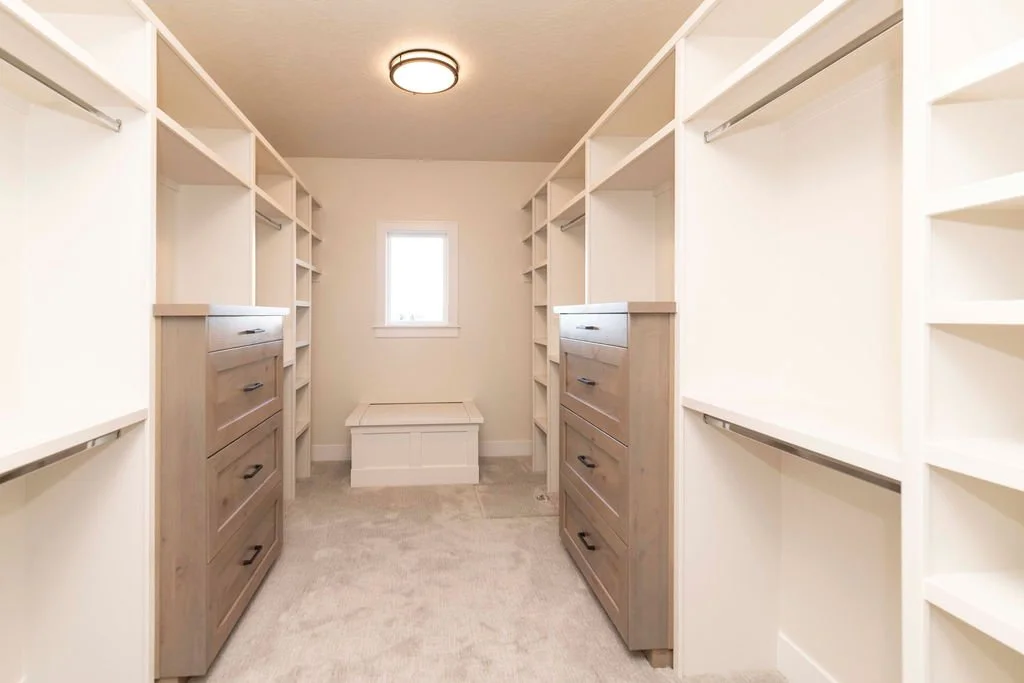 Empty walk-in closet with built-in shelves, drawers, and hanging rods, illuminated by ceiling light and natural light from a small window.