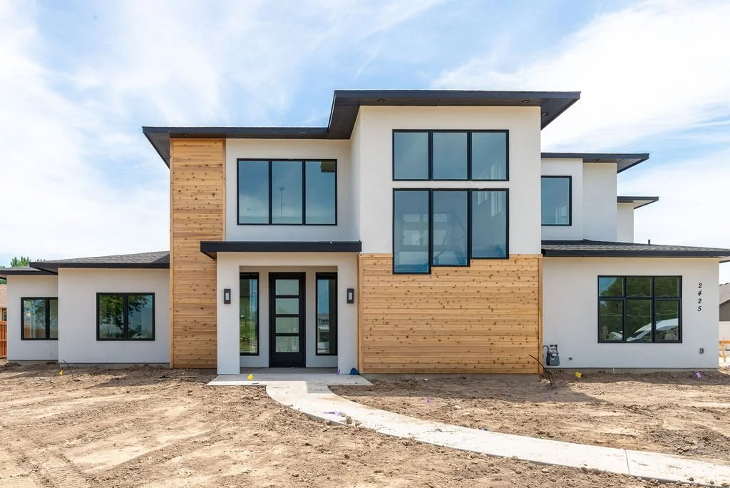 Modern two-story house under construction with white walls, large black-framed windows, and wooden accents on the facade, situated on a dirt lot.