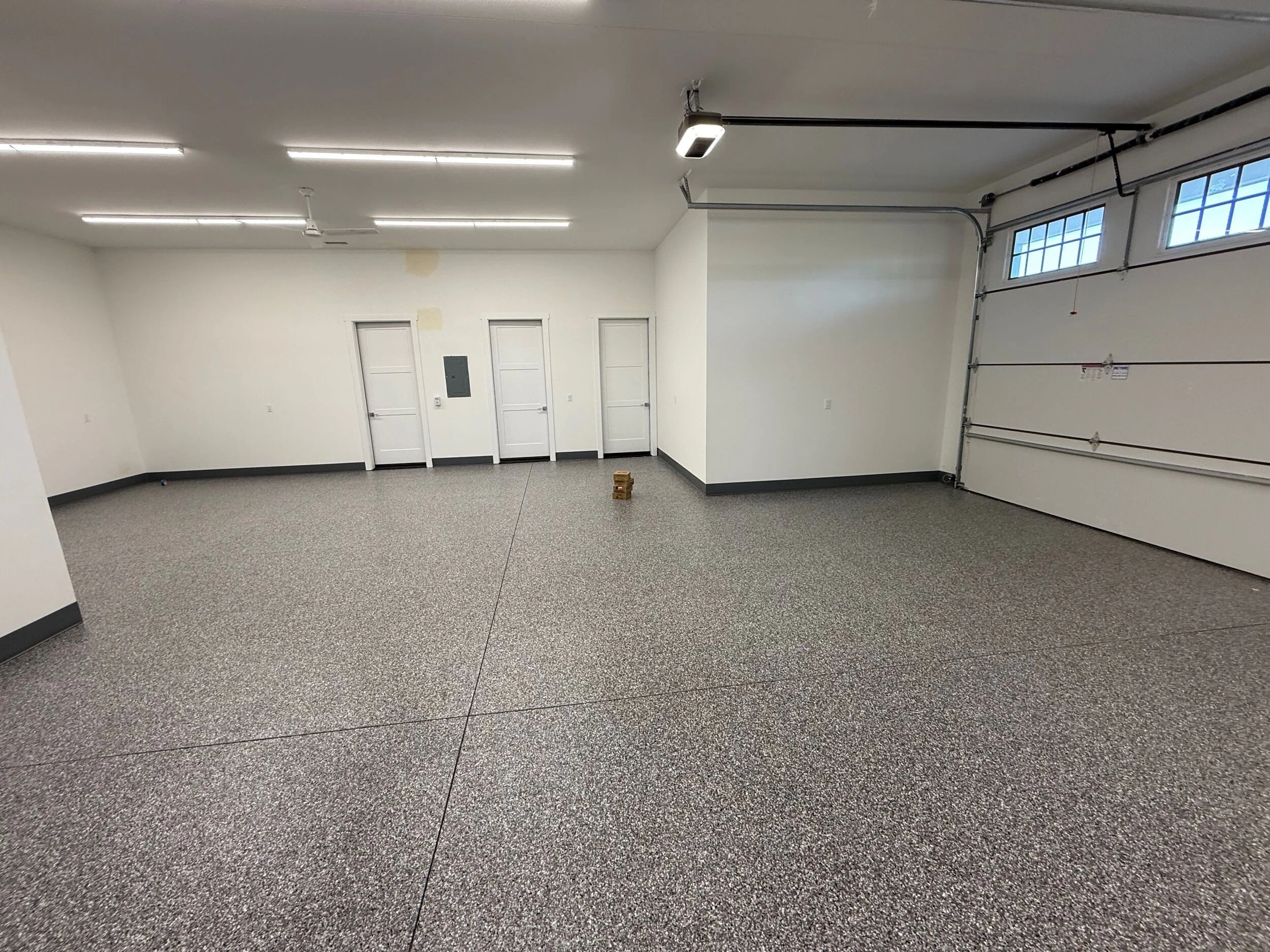 Empty garage with white walls, gray speckled flooring, and a white garage door with windows. Fluorescent lights on the ceiling and a few doors on one wall.
