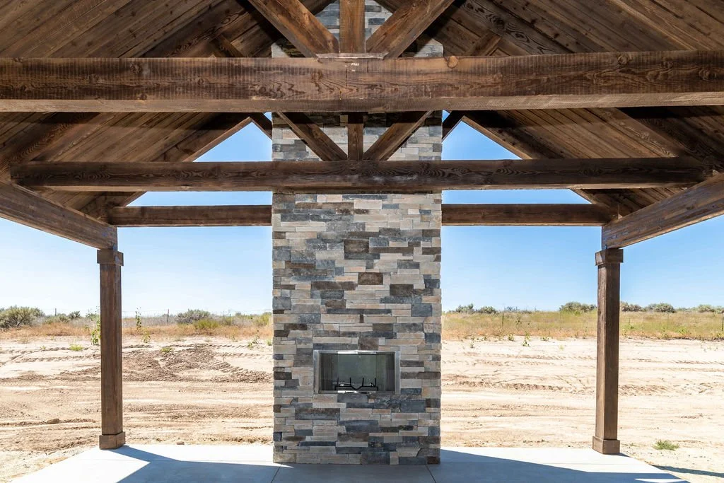 Outdoor structure with a stone fireplace, wooden roof, and open sides, situated in a desert-like landscape.