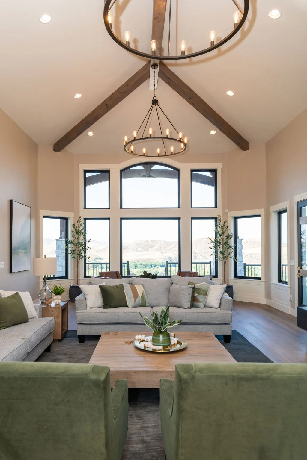 A living room with high vaulted ceiling featuring exposed wooden beams and a large round chandelier. The room has tall windows with a view of mountains and includes two white sofas with pillows, green armchairs, a wooden coffee table with plants, and