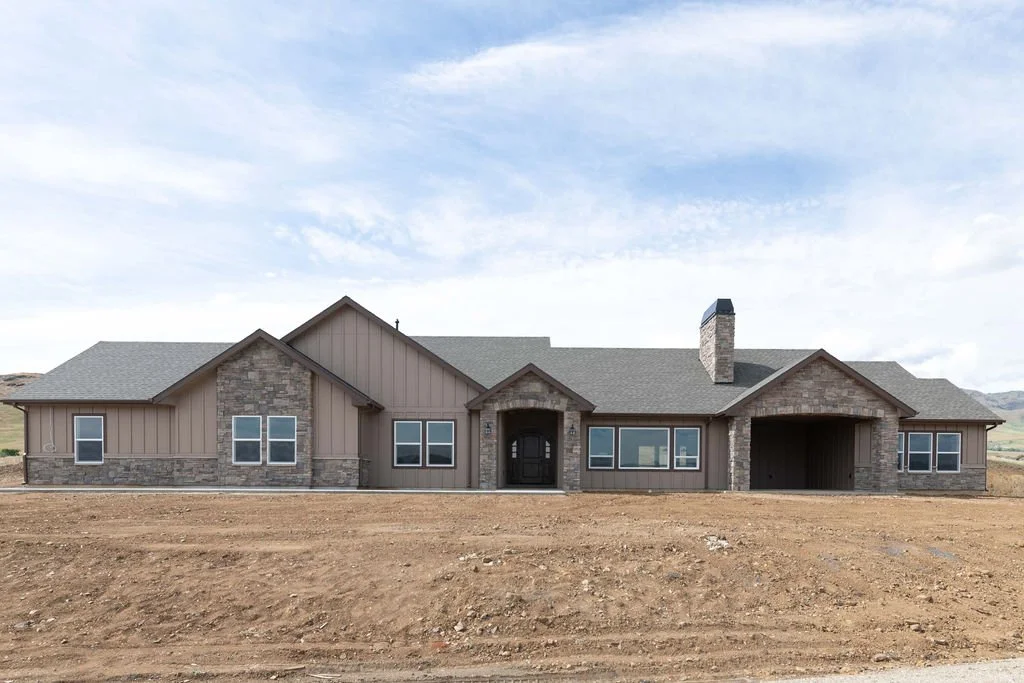 Newly built house with stone and wood siding, multiple windows, and a pitched roof, set in a barren dirt landscape under a partly cloudy sky.