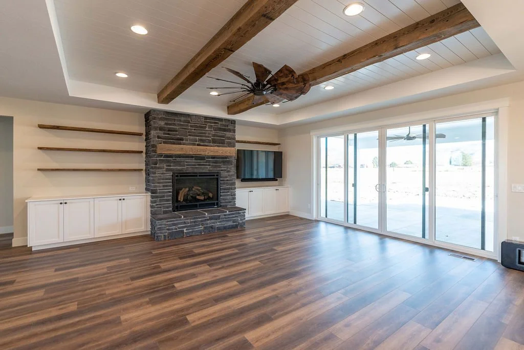 Living room with wood flooring, a stone fireplace, built-in white cabinets, open wooden shelves, a flat-screen TV, large glass sliding doors, and a ceiling fan with wooden beams.