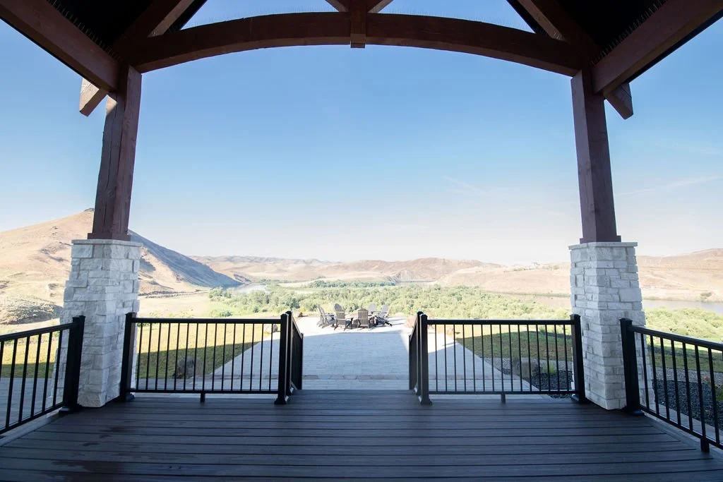 A view from a covered deck overlooking a landscape with hills, green trees, and a clear blue sky, with outdoor chairs in the distance.