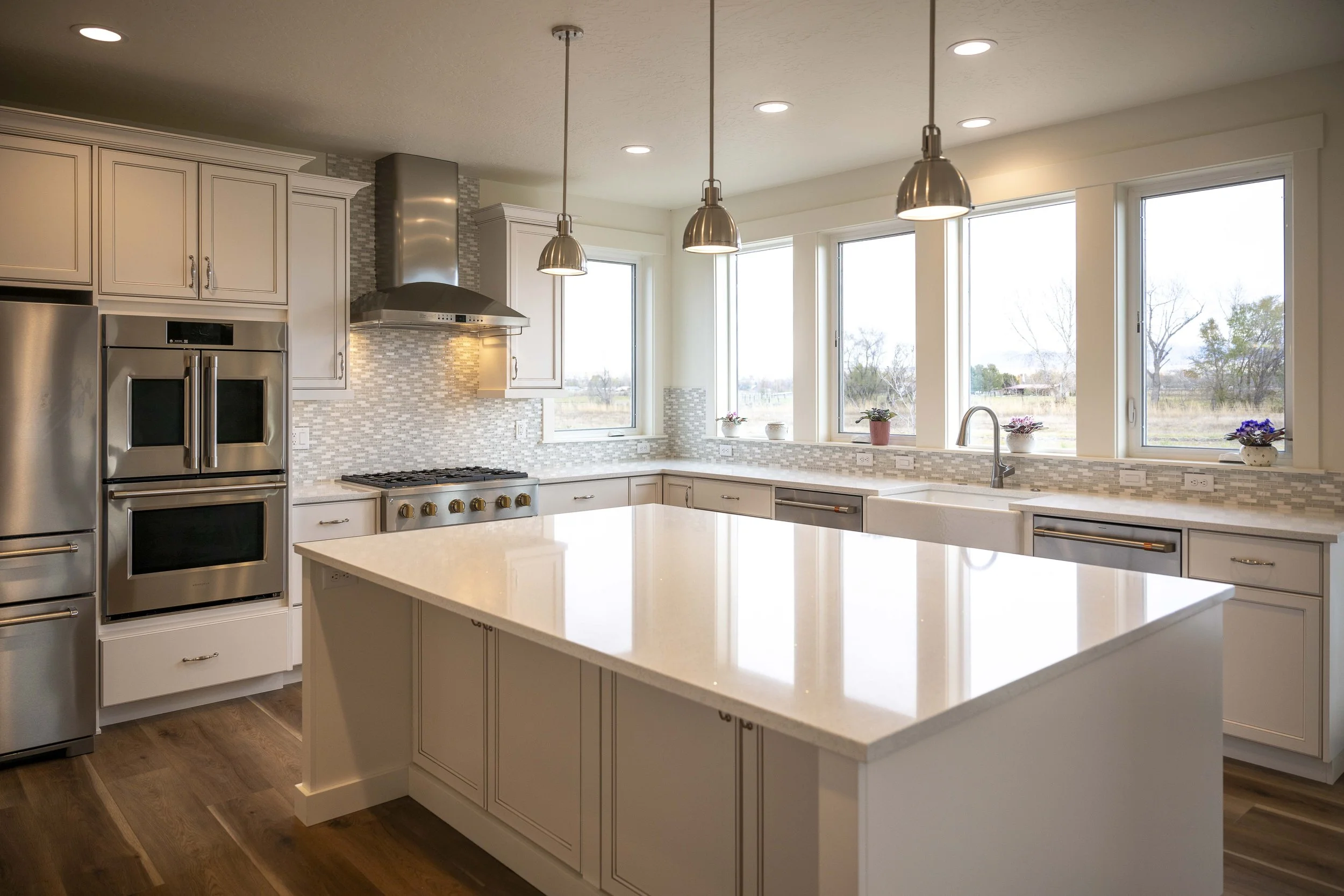 Modern kitchen with white cabinets, stainless steel appliances, a large island with a white countertop, a stainless steel range hood above the stove, large windows three small potted plants on window sills, pendant lights hanging from the ceiling, an