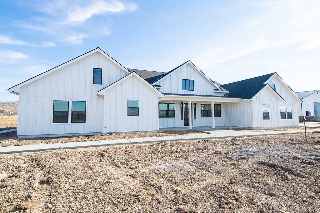 Newly constructed white house with multiple gabled roofs under a blue sky, front yard is unfinished with dirt and a concrete sidewalk.