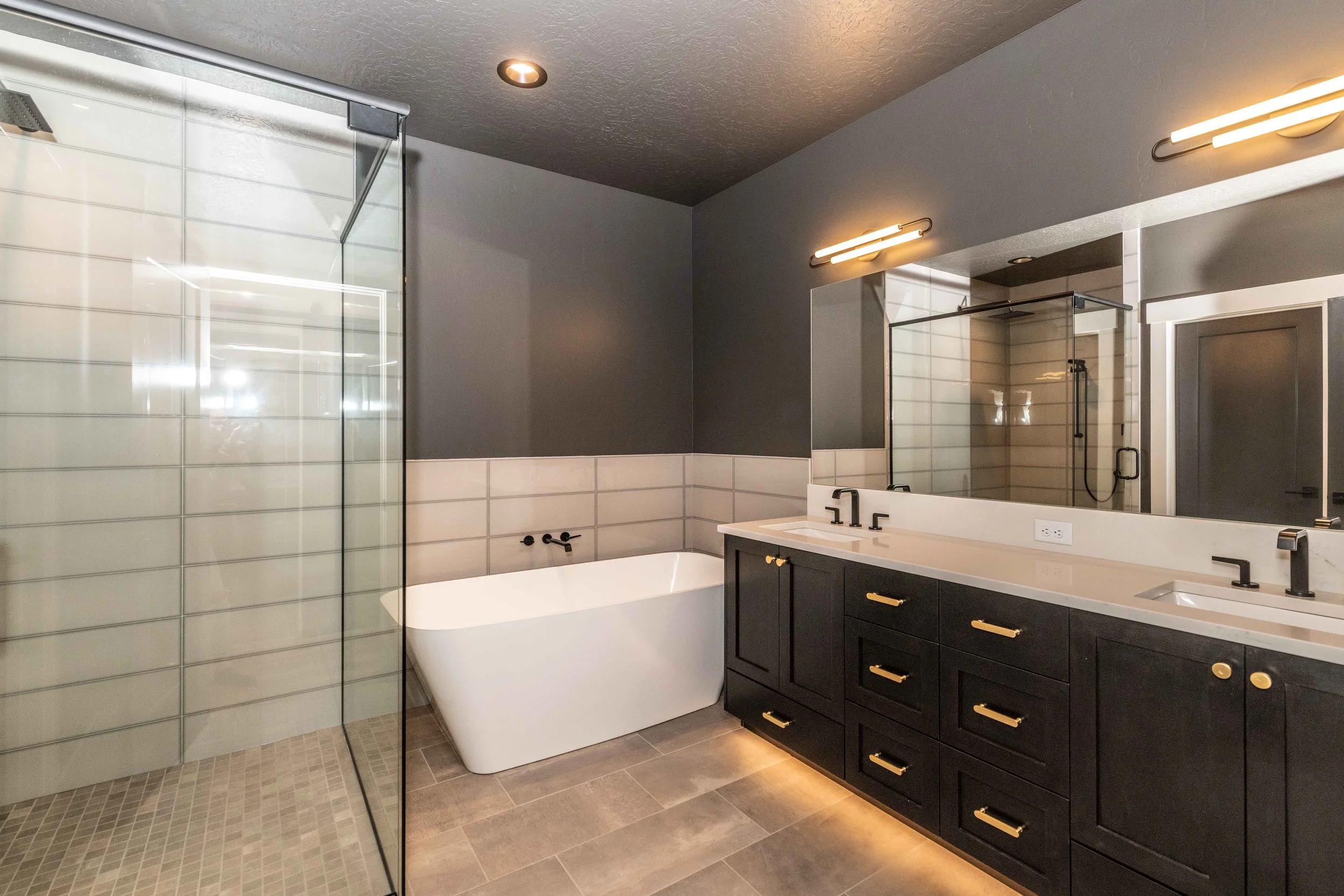 Modern bathroom with a large mirror, dual sinks with dark cabinetry, a bathtub, and a glass-enclosed shower with tiled walls.