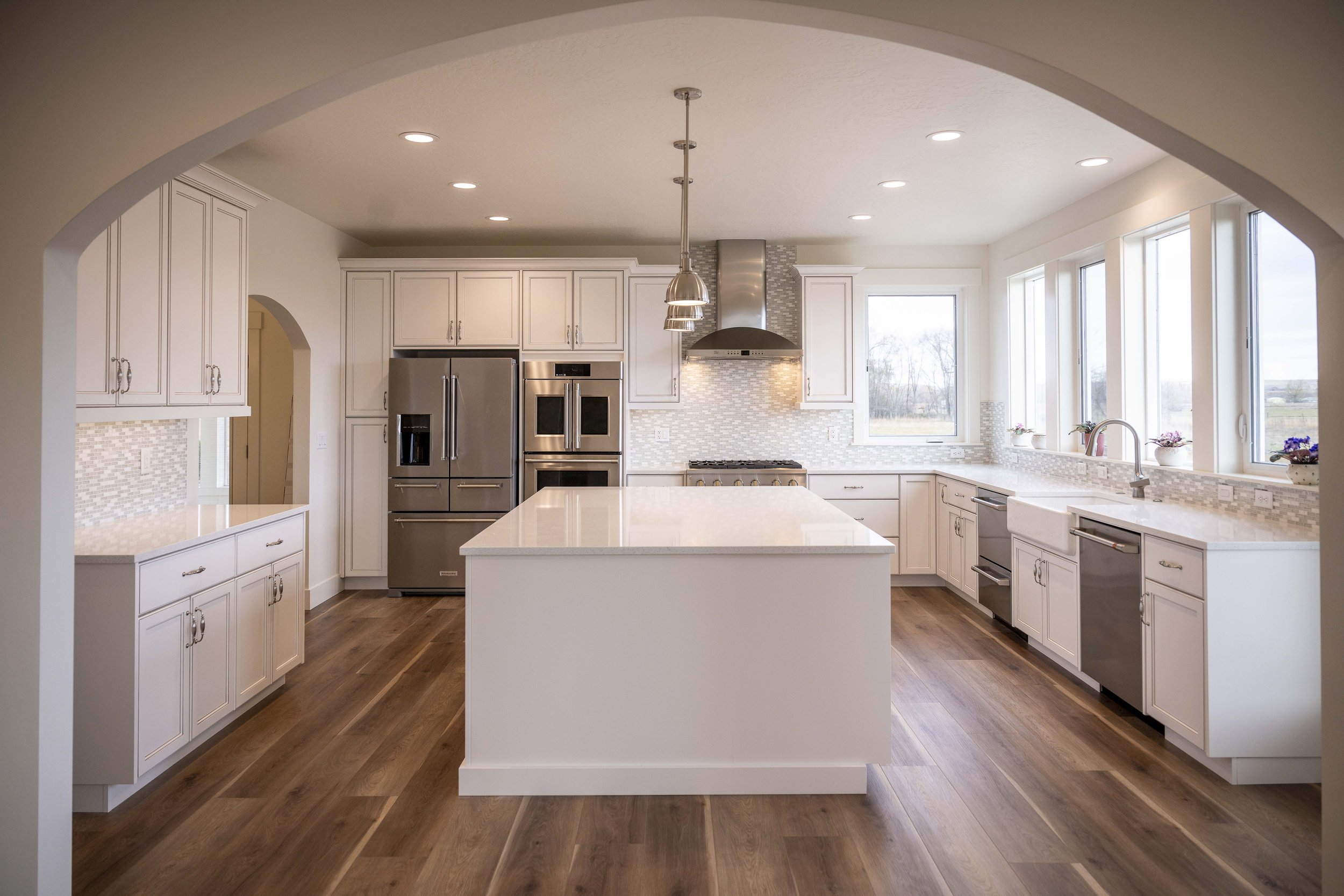 A modern kitchen with white cabinets, stainless steel appliances, hardwood floors, large windows, and pendant lighting.
