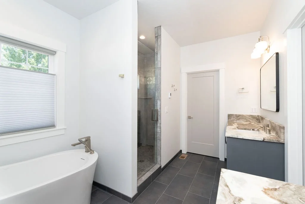 Modern bathroom with a white bathtub under a window, glass shower stall, gray vanity with marble countertop, and a wall-mounted mirror.