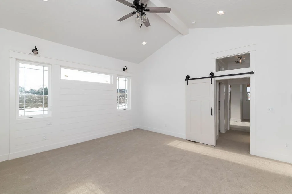Empty bedroom with white walls, beige carpet, ceiling fan, sliding barn door, and windows.
