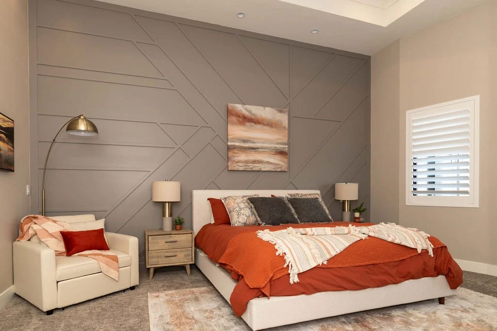 A bedroom with a white bed, orange bedding, and grey accent pillows. The wall behind the bed has a geometric pattern and a landscape painting. There are two nightstands with lamps, a beige armchair with a red pillow and pink blanket, and a window wit