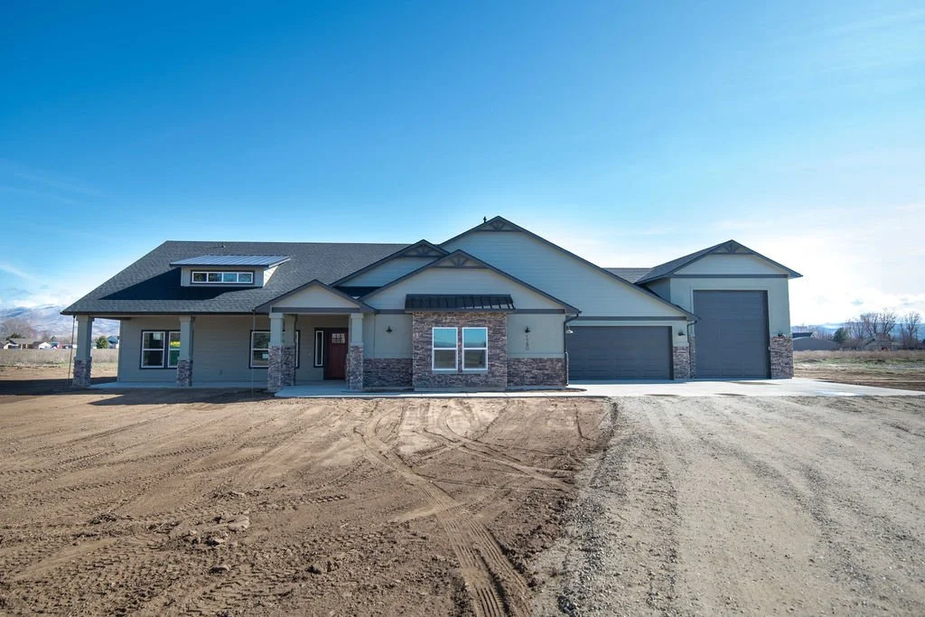 Newly constructed single-family house with light gray siding and stone accents, two-car garage, on a dirt lot under a clear blue sky.