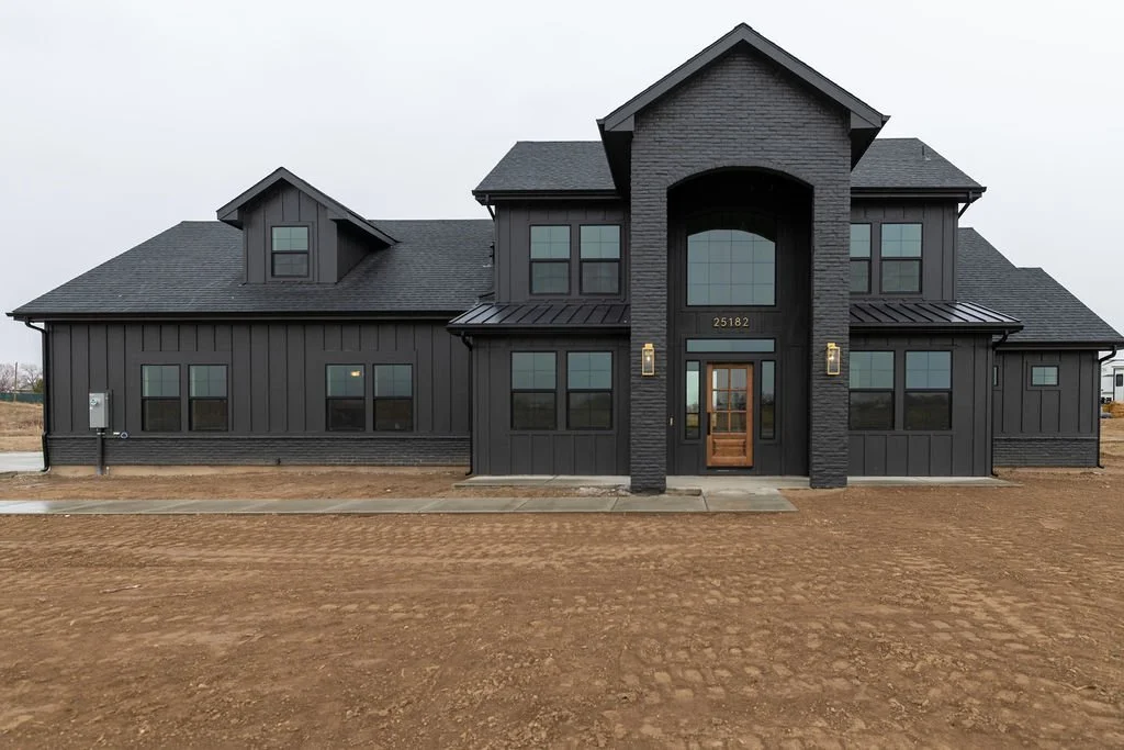 A modern two-story house with a dark exterior, large front entrance, and multiple windows, set on a plot of bare, dirt land.