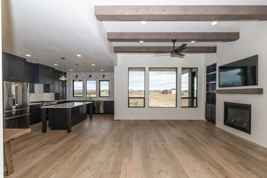 Open-concept living room and kitchen with large windows, wood ceiling beams, a ceiling fan, mounted TV, and a gas fireplace.