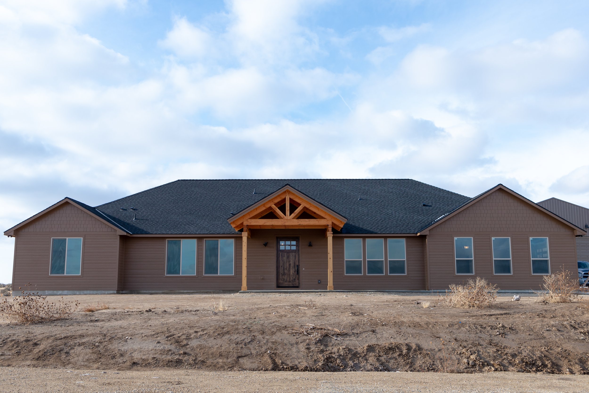A house under construction or renovation with a newly built front porch, multiple large windows, and a dark-shingled roof. The yard is bare dirt with some weeds and bushes, and the sky is partly cloudy.