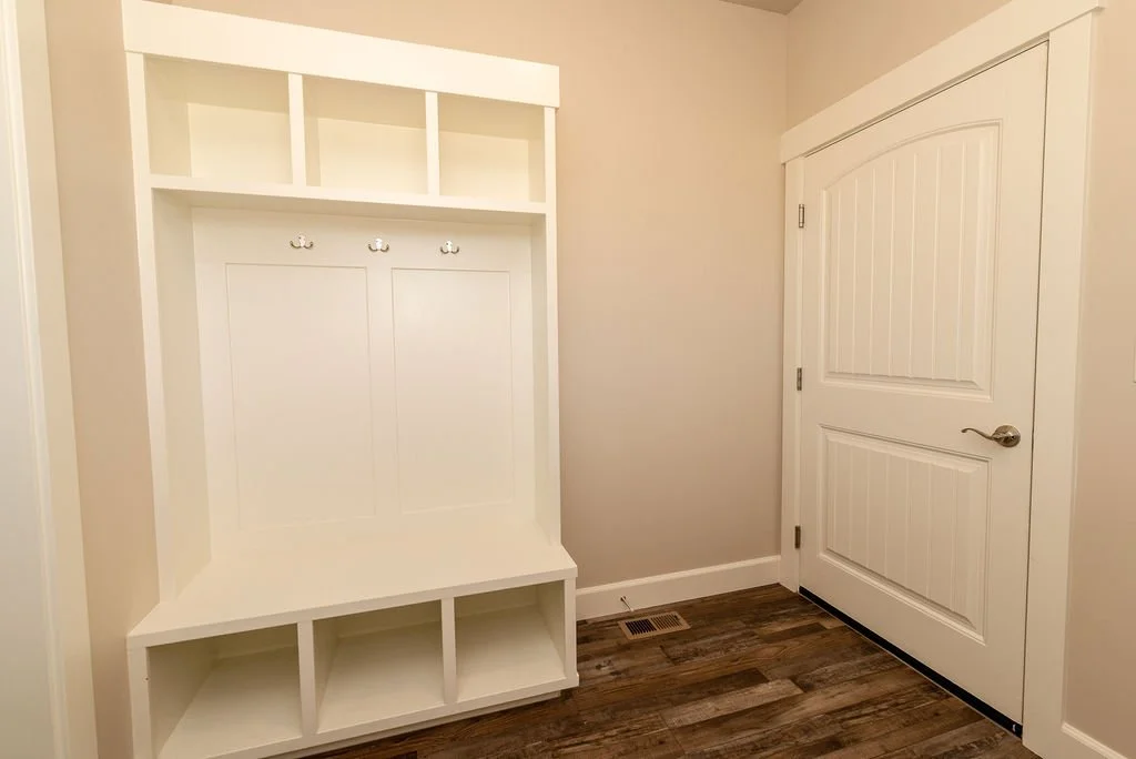 White hall coat rack with hooks and storage compartments next to a closed white door on a wooden floor.