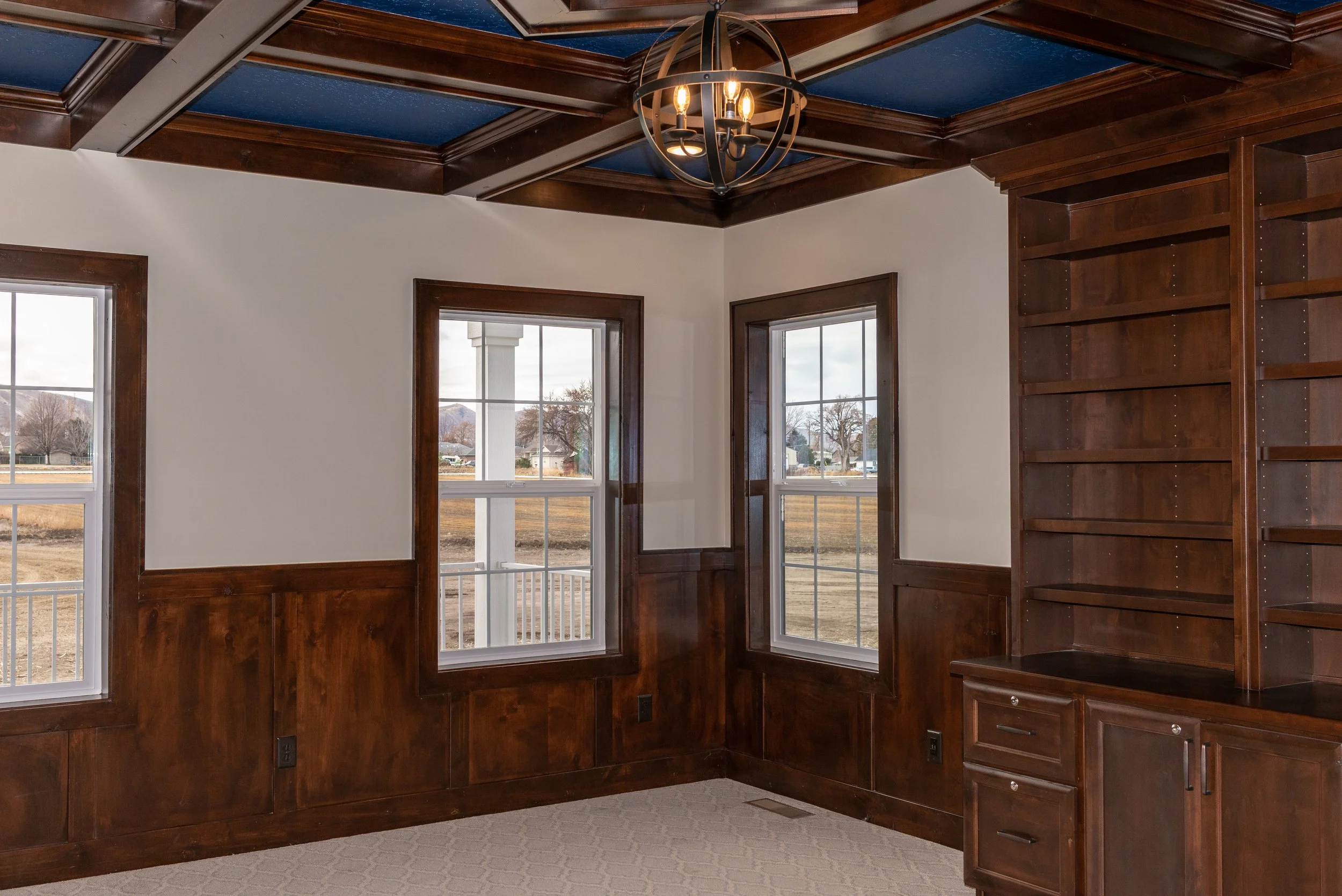 Interior of a room with dark wooden trim, crown molding, and a coffered ceiling painted blue. There are three large windows with white frames showing an outdoor view of trees and a street. A black metal chandelier with five bulbs hangs from the ceili