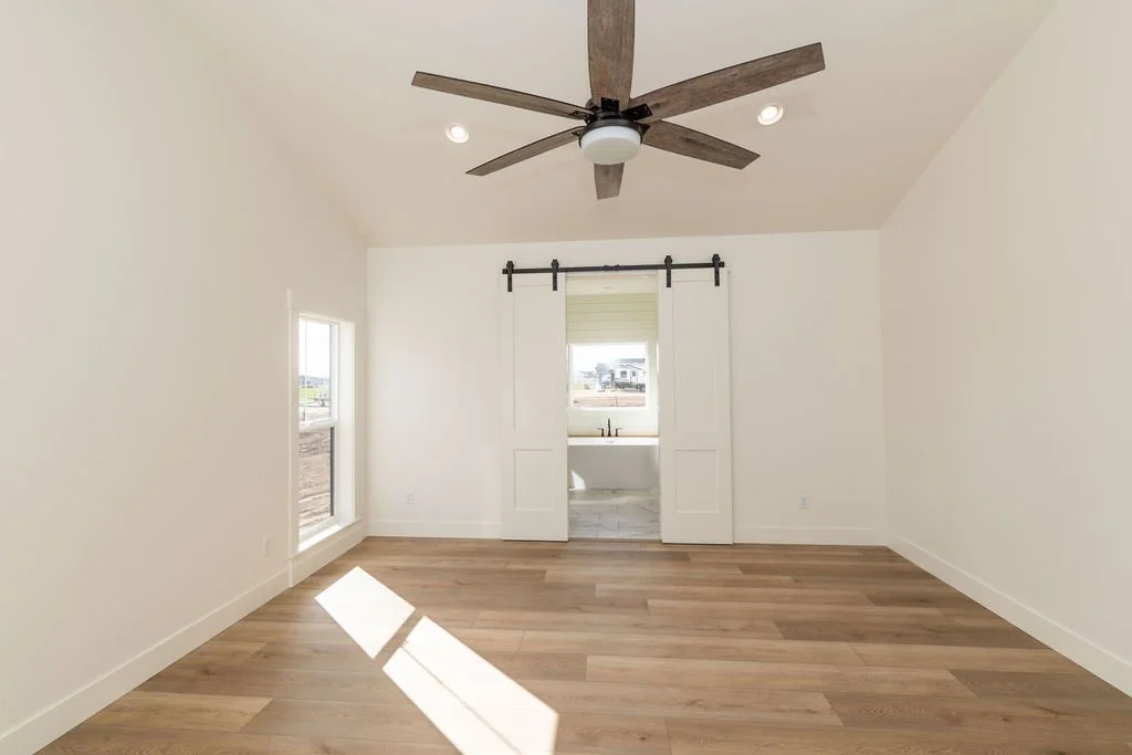 Empty room with wooden floor, two windows, a ceiling fan, and sliding barn doors leading to a bathroom.