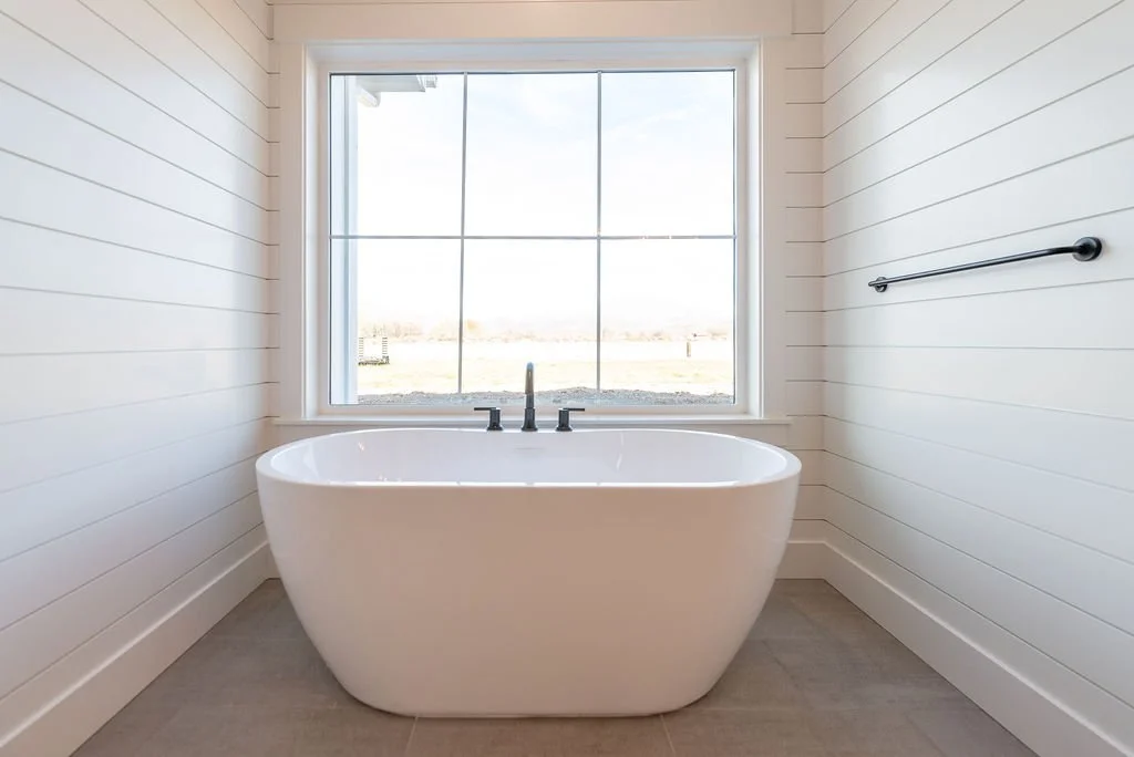 White freestanding bathtub in a bright bathroom with white shiplap walls and a large window showing an outdoor view.