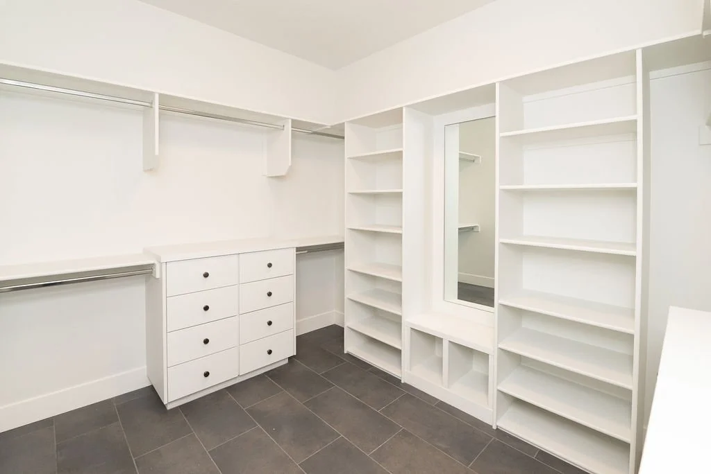 Empty walk-in closet with white shelving, drawers, mirrored door, and dark tile floor.
