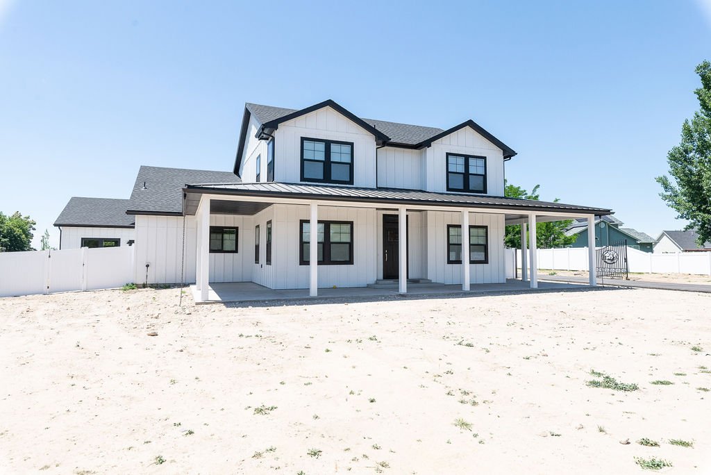 A modern white two-story house with black window frames and a covered front porch, situated on a sandy lot with a white fence and clear blue sky.
