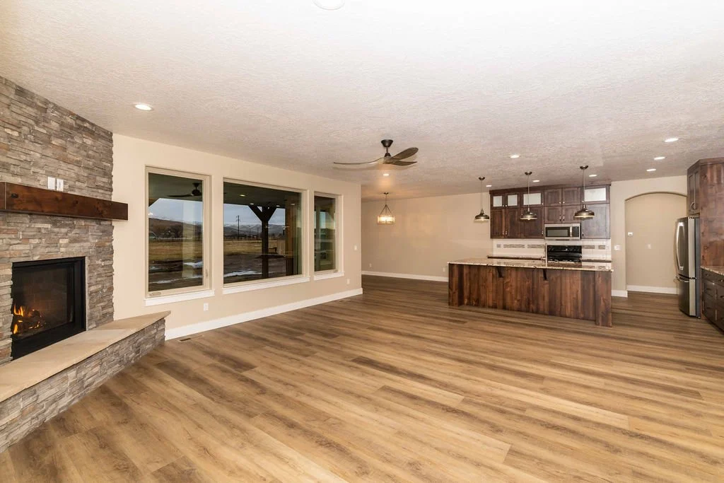 Spacious open-concept living room and kitchen with hardwood floors, stone fireplace, large windows, and modern kitchen with wooden cabinets and island.