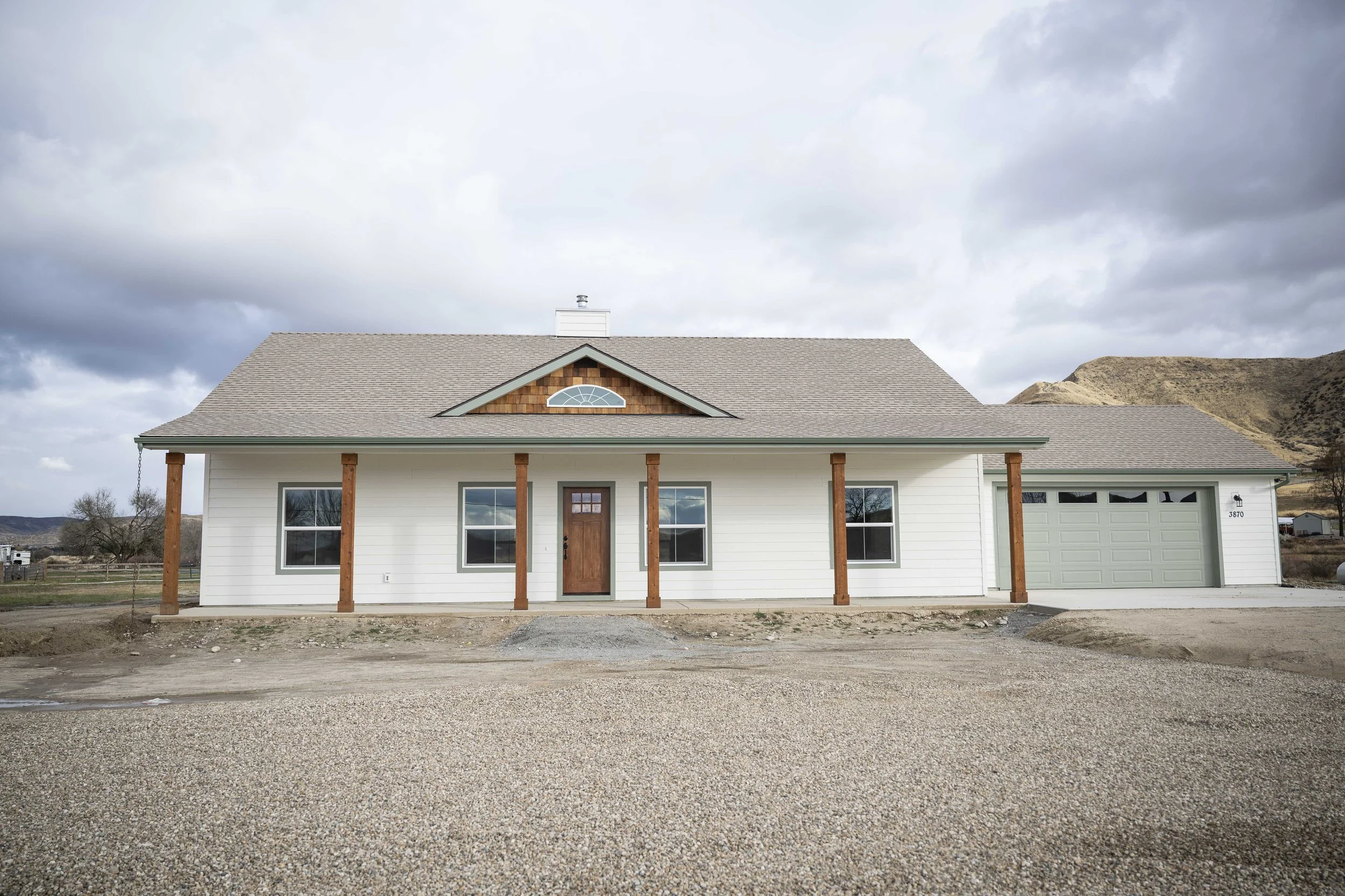 New white house with a brown front door, two large windows, and a garage, set against a cloudy sky and mountainous landscape.