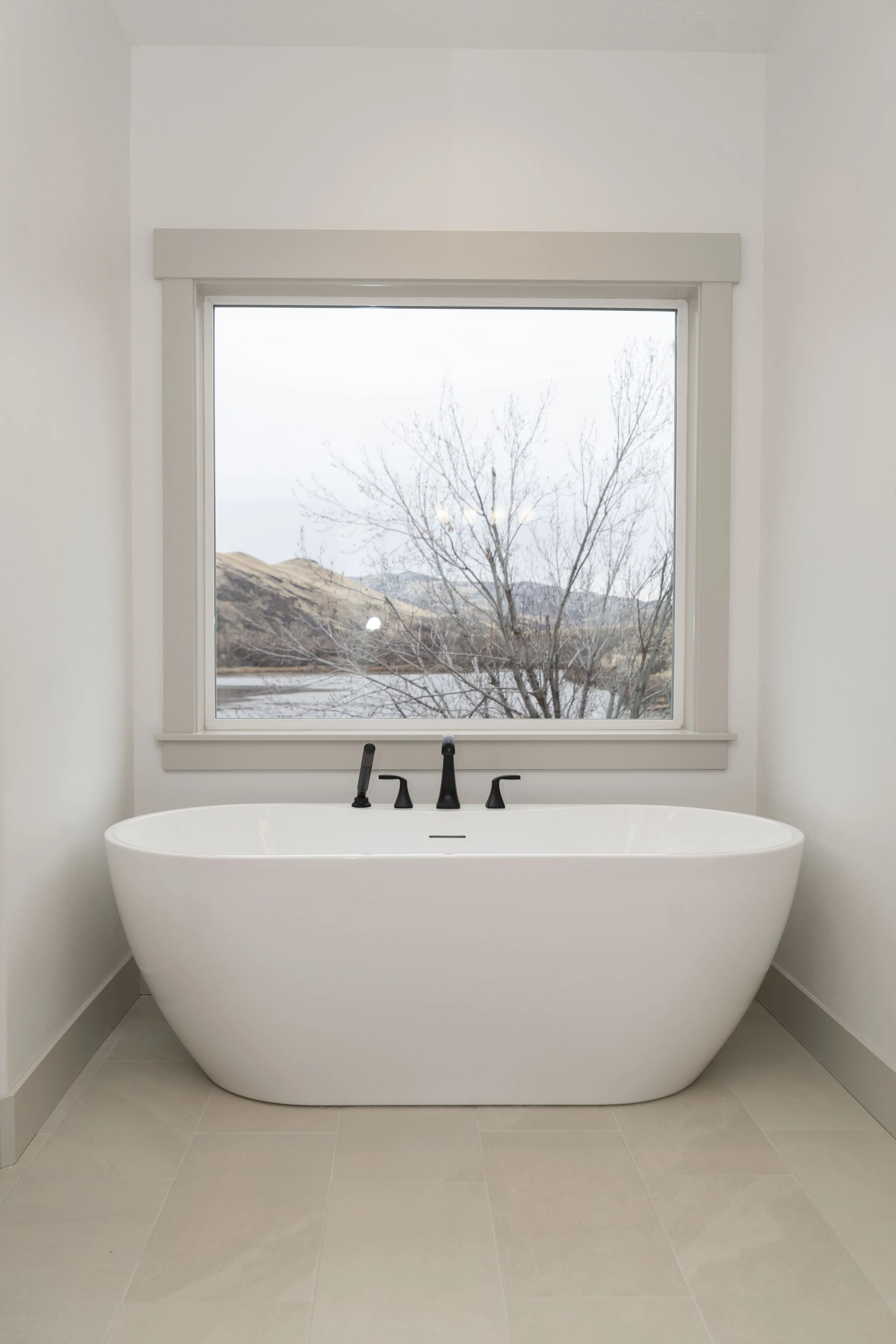 Bathroom with a freestanding white bathtub, black faucet, and a large window showing a view of barren trees and hills.