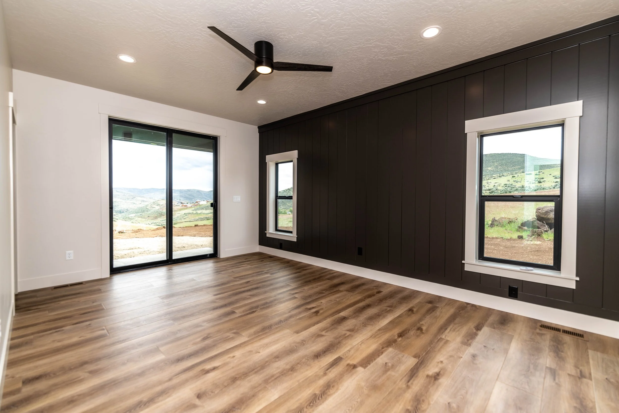 An empty room with hardwood floors, a black accent wall with two windows, and a sliding glass door. The room has a ceiling fan with three black blades, recessed lighting, and large views of a green, mountainous landscape outside.