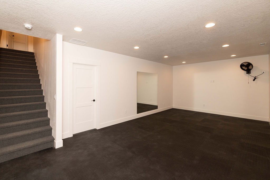 Empty basement room with white walls, dark carpeted floor, and stairs leading up on the left. There is a small door and a mirrored opening on the wall, with a wall-mounted fan in the top right corner.