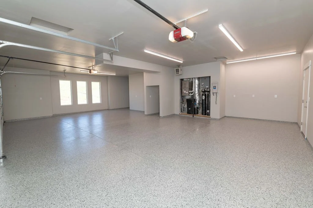 Empty spacious garage with epoxy flooring, white walls, ceiling-mounted garage door openers, windows, and utility panel.