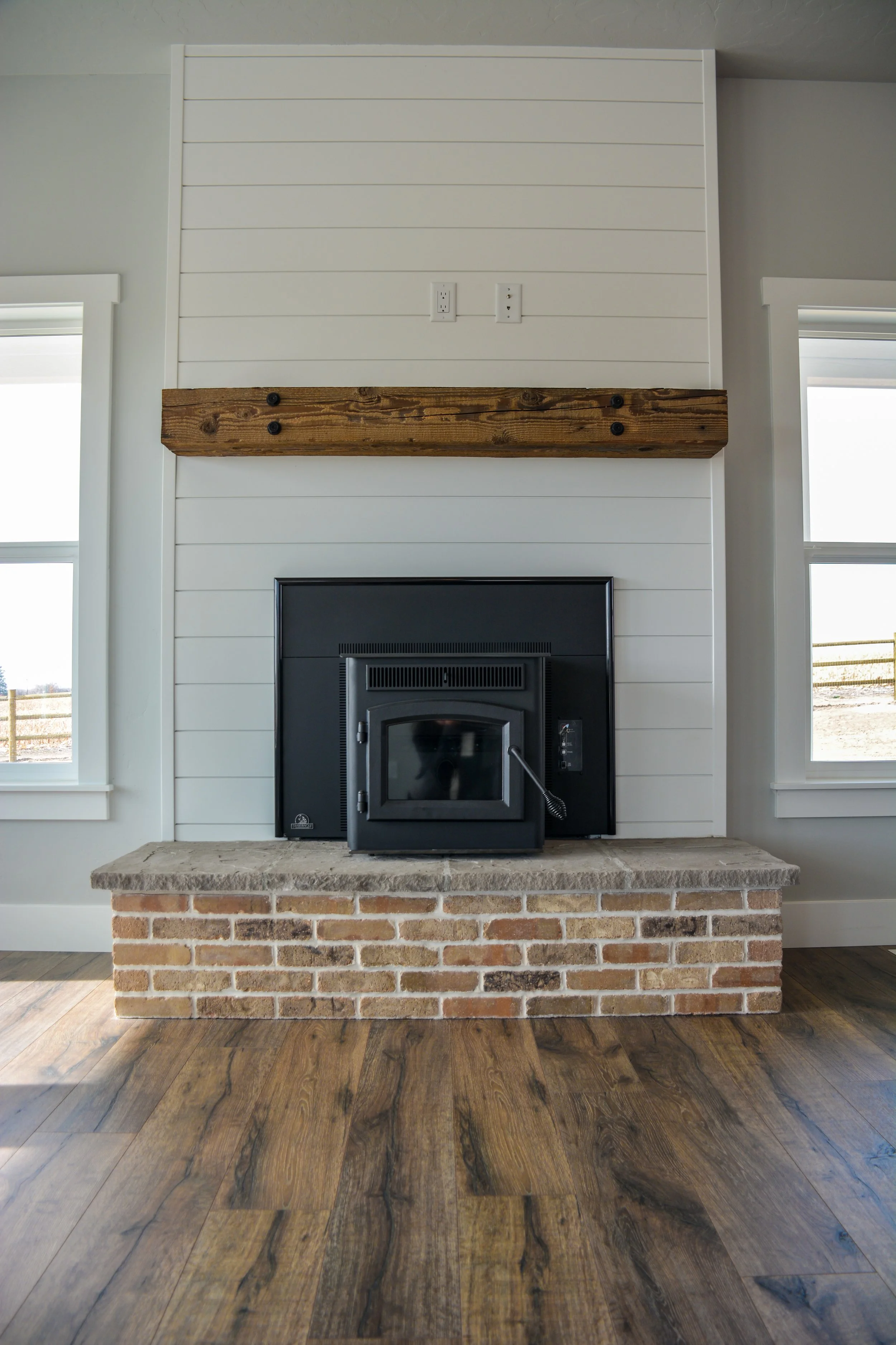 A fireplace with a black pellet stove insert, mounted on a brick hearth, with a wooden mantel and white shiplap wall behind. Two windows flanking the fireplace, with outdoor views.