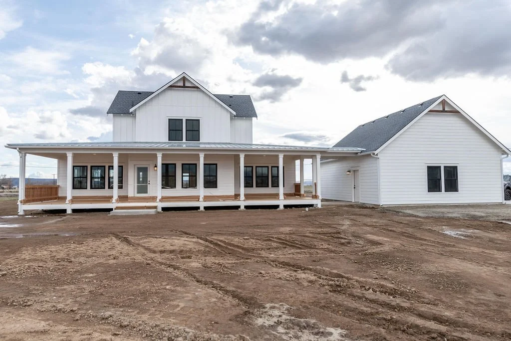 Newly built white house with a large front porch, located in a dirt lot, under a cloudy sky.
