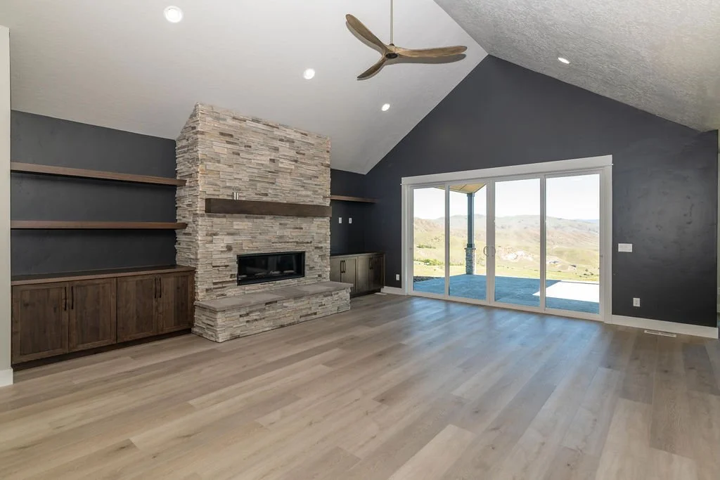 Living room with a stone fireplace, dark blue accent wall, built-in wooden cabinets and shelves, large sliding glass doors showing outdoor landscape, ceiling fan, hardwood flooring.