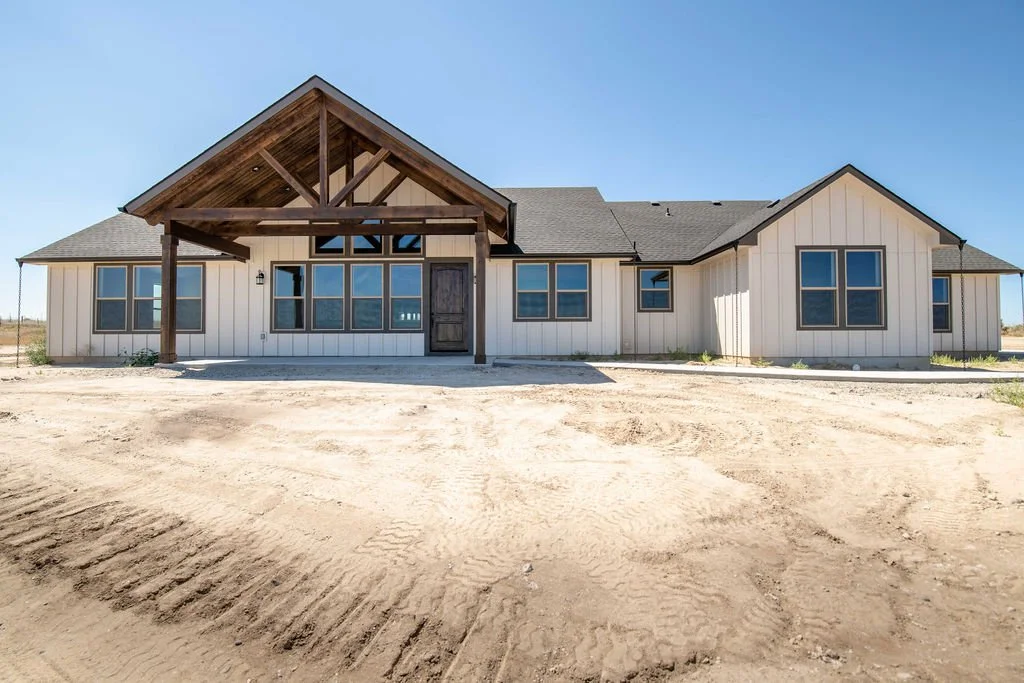 Newly built single-story house with white siding, multiple windows, front porch, and gabled roof, located in an area with dirt ground and clear blue sky.