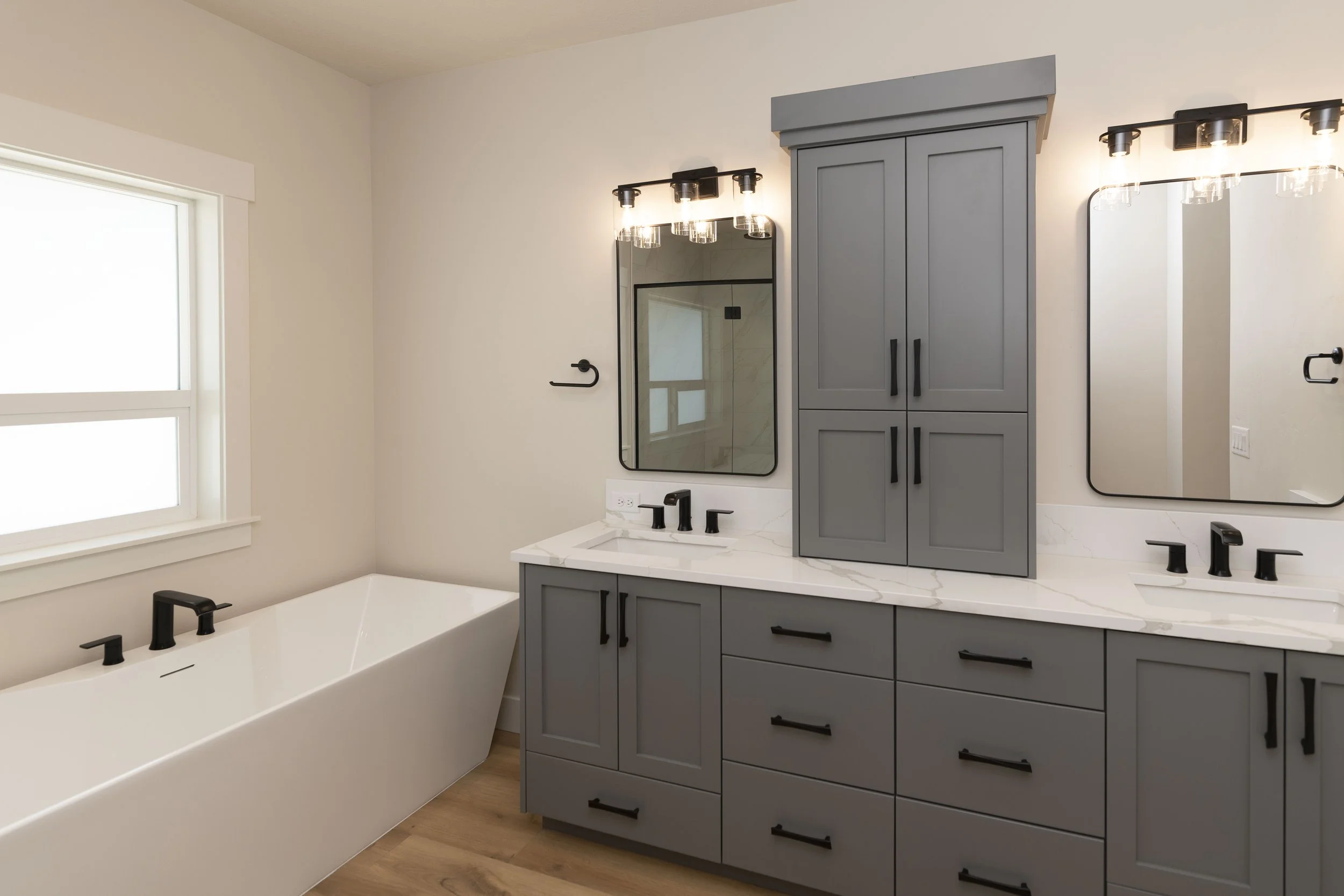 A modern bathroom with a gray cabinet, two mirrors with black frames, black fixtures, and a bathtub, illuminated by wall-mounted lights.