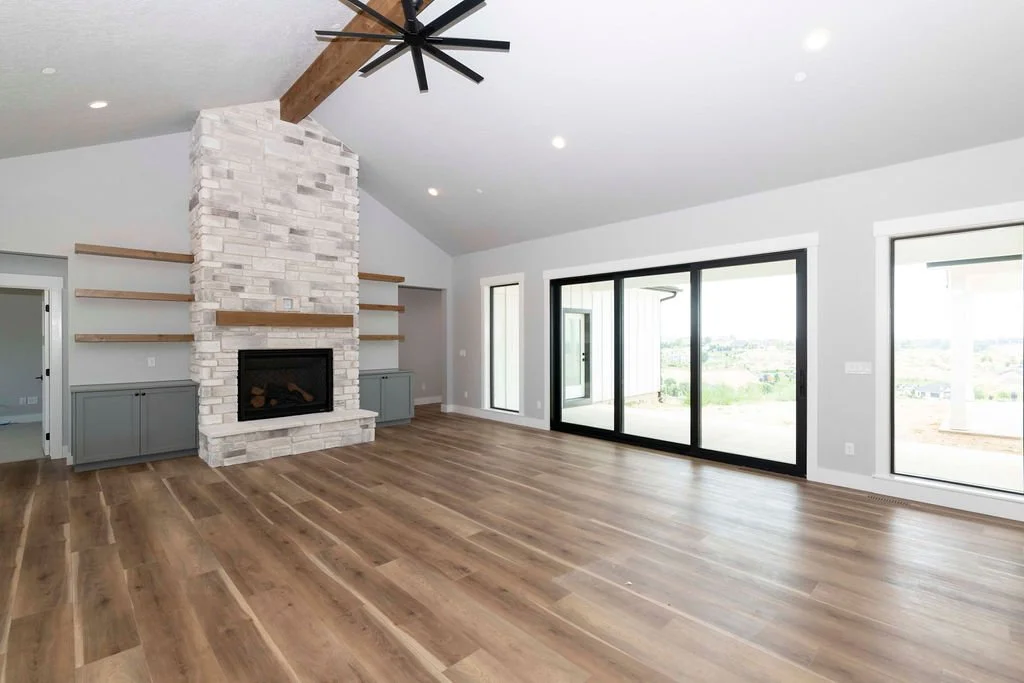 Empty living room with a fireplace, wooden floors, large sliding glass doors, and windows showing an outdoor view.