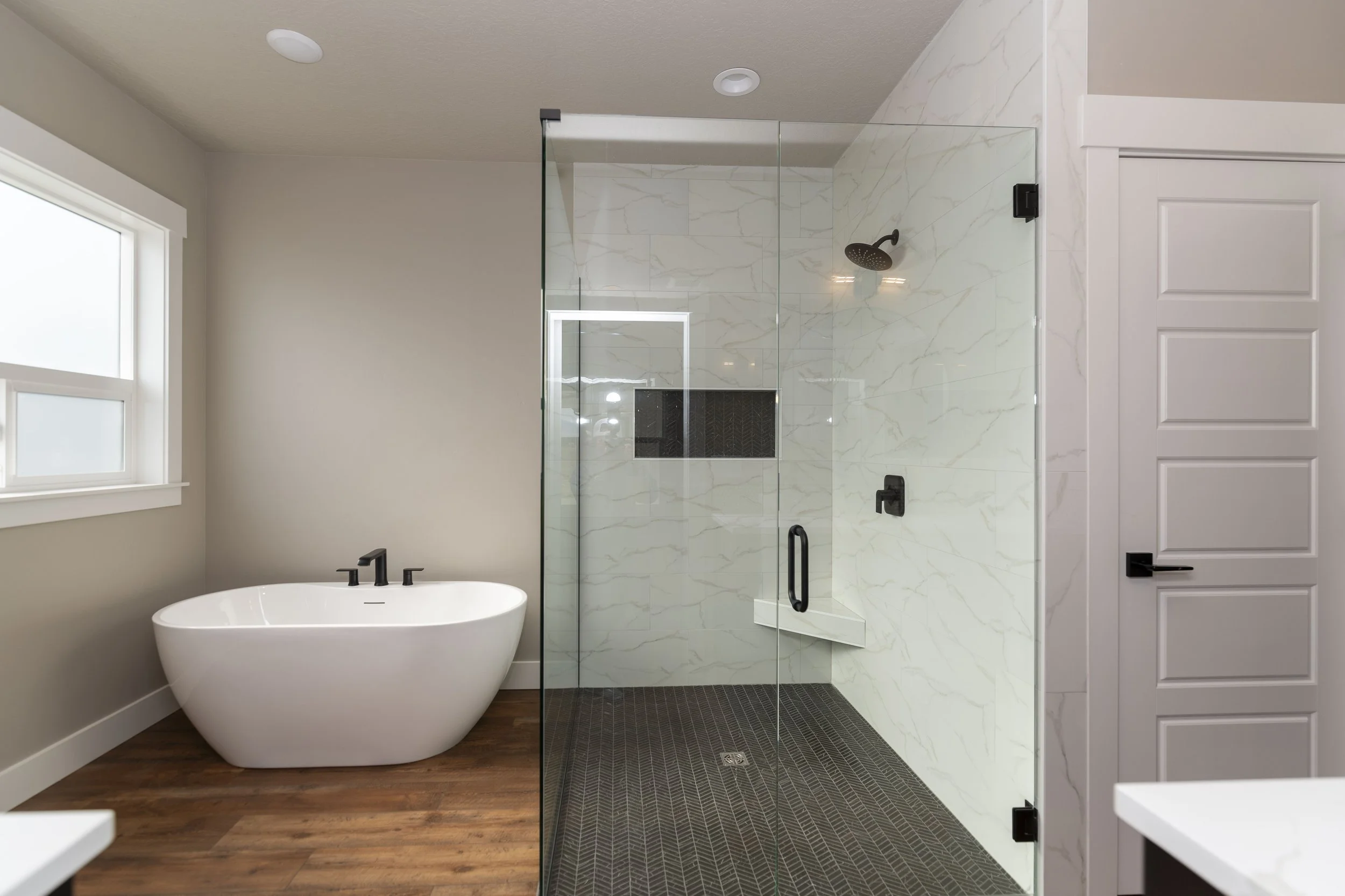 A modern bathroom with a freestanding bathtub, a glass-walled shower with marble tiles, a window, and wood flooring.
