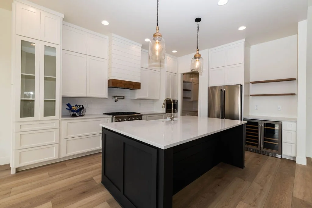 Modern kitchen with white cabinets, a large dark island with a white countertop, pendant lights, and stainless steel appliances, including a refrigerator and wine cooler.