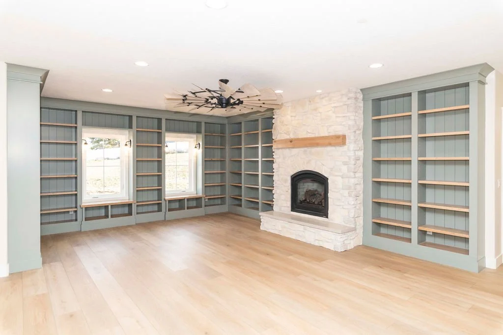 Empty living room with built-in bookshelves, a white stone fireplace, light hardwood flooring, and two windows with outdoor views.