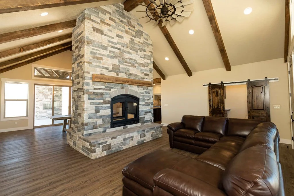 Living room with a brown leather sectional sofa, a stone fireplace, wooden beams on the ceiling, and large windows with a sliding door leading outside.