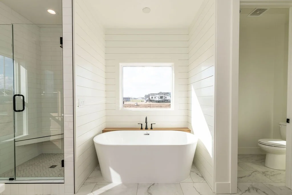 Modern bathroom with white shiplap walls, a large window, a freestanding bathtub beneath the window, and a glass-enclosed shower on the left. A toilet is visible in the adjacent room.