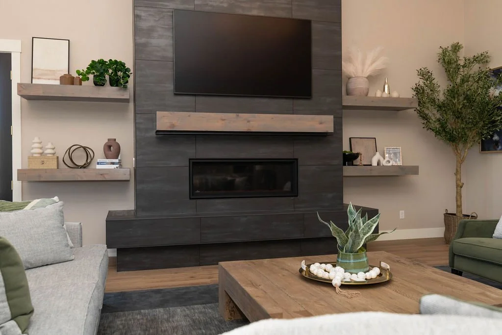 Living room with a gray sofa, wooden coffee table, and a black fireplace with a mounted TV above it. Open shelving with decorative items and plants, a large potted plant in the corner.