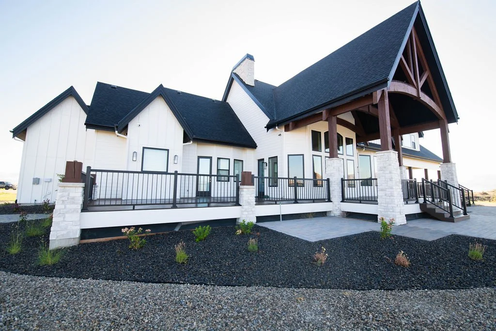A modern house with white exterior walls, large windows, a black roof, and a spacious deck with black railings, stone pillars, and steps leading to the ground, surrounded by gravel landscaping.
