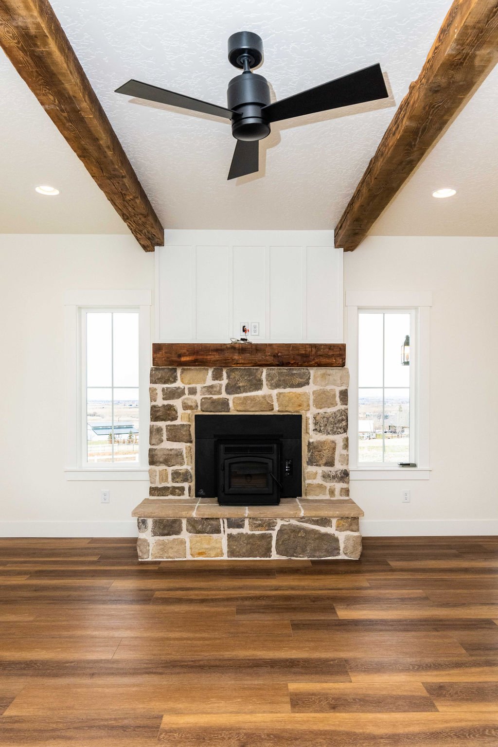 Living room with a stone fireplace, white walls, wooden floor, and a ceiling fan with black blades.