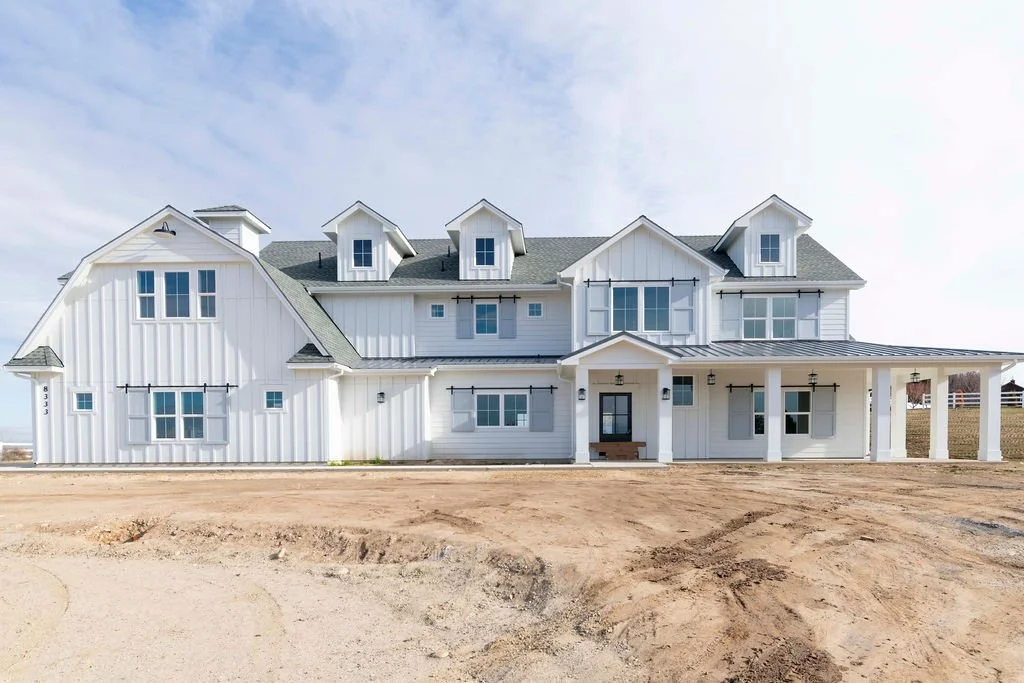 Newly constructed white farmhouse-style house with gray roof, front porch, multiple windows, and three dormer windows on the roof, set on a dirt lot.