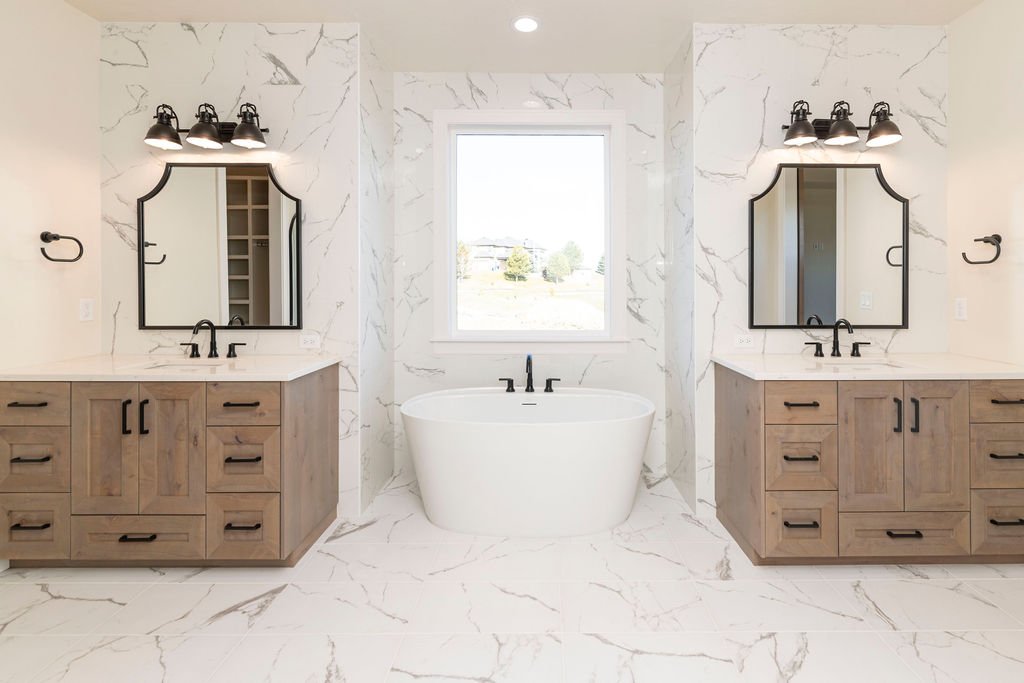 Modern bathroom with a white marble floor and walls, two wooden vanities with black handles and mirrors, black light fixtures above each mirror, a freestanding bathtub below a window in the center.