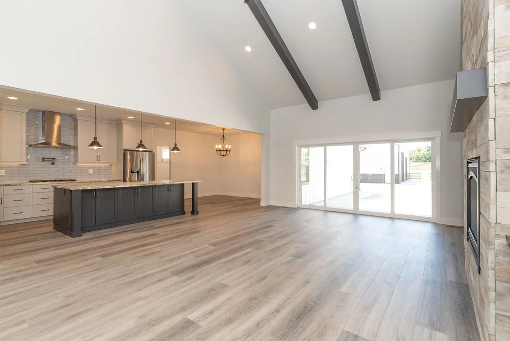 Empty open-concept living room and kitchen with high vaulted ceiling, wood flooring, a large sliding glass door, and a stone fireplace.